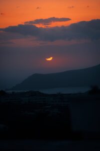 Sunset over hills with orange clouds, silhouetted buildings in foreground, tranquil setting.