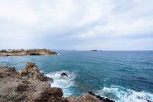 Rocky cliffs and blue ocean with waves, distant rock formations, and coastal buildings.