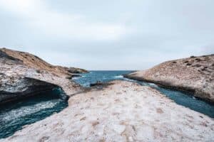 Rocky coastal landscape with a natural arch, turquoise water, and overcast sky.