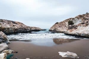 Secluded beach with narrow inlet, rocky hillsides, overcast sky, waves on sandy shore.