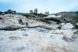 Rocky coastal landscape with natural pool, white building, trees, and hills under clear sky.