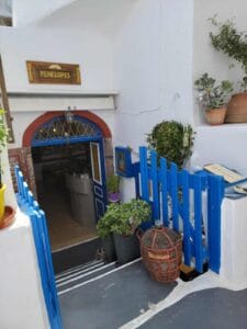 White building with blue entrance, sign "Penelopes", potted plants, wicker bottle, blue fence.
