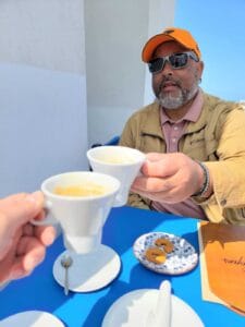Person in sunglasses and orange cap at blue table holding coffee, sharing moment with another.