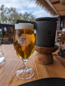 Glass of beer with foamy head on outdoor wooden table, decorative logo, blurred trees behind.