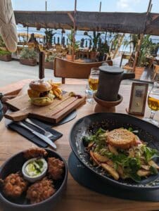 Outdoor dining setup with burger, fries, salad, fried bites, beer, sea view, and seating.