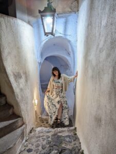Person in floral outfit in narrow, curved alley with textured walls, cobblestone path, under lantern.