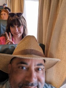 Man in straw hat smiling on train, woman behind makes peace sign, beige curtains and seats.