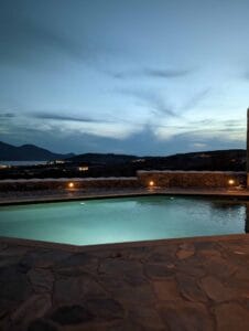 Illuminated pool on stone patio, low wall, evening sky, distant hills in twilight.