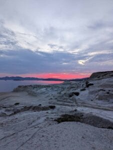 Coastal landscape at sunset with rocky terrain and pink horizon; islands in calm sea.