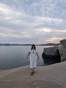 Person in white dress and sandals stands on rocky ledge by coastal cliffs and calm water.