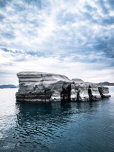 Rock formation with layered patterns rises from the blue sea under a cloudy sky, distant hills visible.