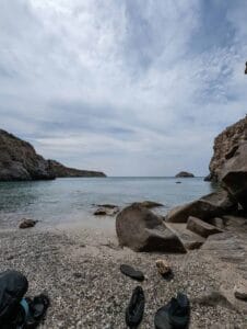 Rocky beach with calm water, cloudy sky, cliffs, sandals, and a bag on the shore.