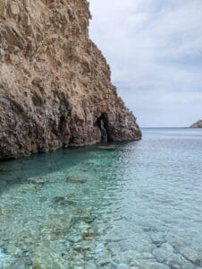 Rocky coast with turquoise water, cliffside cave, overcast sky, sea extending to horizon.