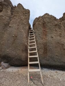 Wooden ladder leans against a rugged rock formation under an overcast sky in a natural setting.