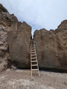 Wooden ladder against a narrow gap in a rocky cliff under an overcast sky.