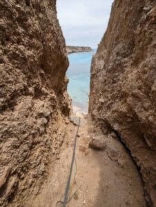 Narrow rocky path between cliffs leading to turquoise sea under lightly clouded sky.