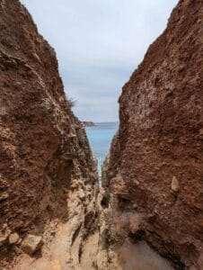 Narrow rocky passage through cliffs leads to the sea under an overcast sky.