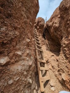 Narrow rocky passage with wooden ladder leading upwards, rugged walls, sky visible above.