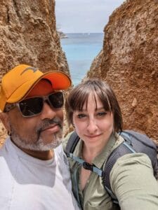 Man and woman take a selfie between cliffs with an ocean view and distant sailboats.