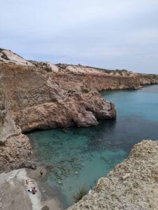 Overcast coastal scene with cliffs, a cove, turquoise water, sandy beach, and people relaxing.