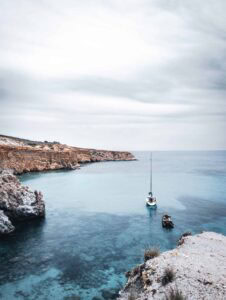 Coastal scene with clear waters, rocky cliffs, anchored sailboat, small vessel, overcast sky.