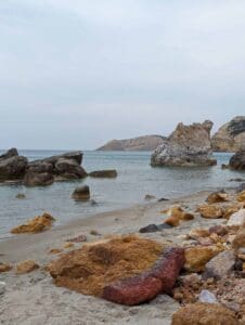 Rocky beach with colorful stones, large formations in clear water, hills in background, cloudy sky.