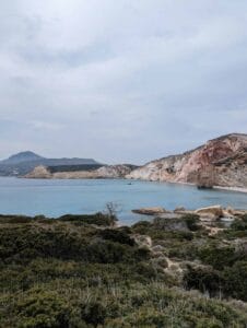 Coastal landscape with blue sea, rocky cliffs, foreground greenery, and cloudy sky.