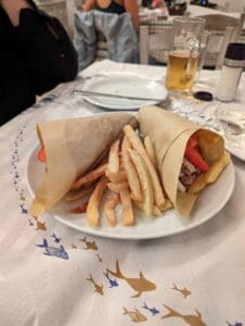 Plate with two wrapped sandwiches, fries, beer, and cutlery on fish-patterned tablecloth.