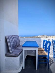Outdoor seating with striped bench, blue table, and chairs; coastal view under blue sky.