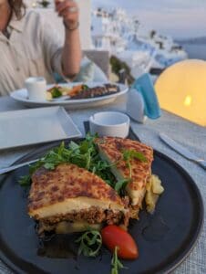 Plate of moussaka with arugula and tomato garnish, person dining, scenic seaside view.
