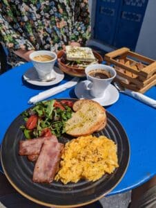 Breakfast with scrambled eggs, bacon, greens, bread, salad, and coffee on a blue table.