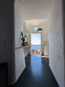 Corridor with sea view, open doorway, round window, small table outside, plant on shelf.