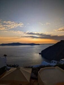Scenic sunset over calm sea with islands, Mediterranean buildings, umbrellas, orange-blue sky.