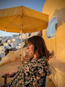 Person in floral outfit on terrace under yellow umbrella, white buildings in sunny background.