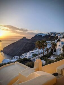 Coastal view with white buildings, blue domes, palm trees, sunset over cliffs, and sea.