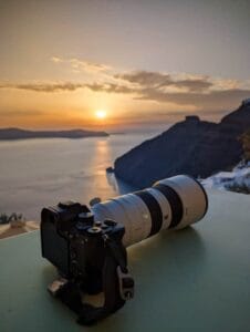 Camera with telephoto lens overlooking a sunset above sea, sky partly cloudy, sun near horizon.
