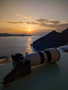 Camera with long lens on surface, sunset over sea, hills silhouetted, warm glow on water.