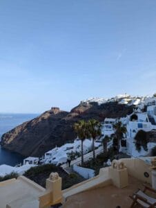 Hillside village with white domed buildings, blue accents, palm trees, cliffs, and ocean view.