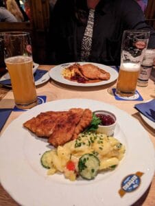 Two plates with schnitzel, potato salad, cranberry sauce, and two glasses of beer on a table.