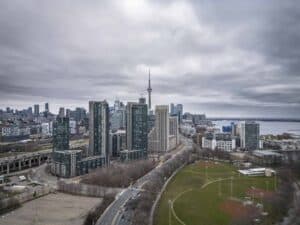 Cityscape with tall buildings, central tower, road, sports field, water, and overcast sky.