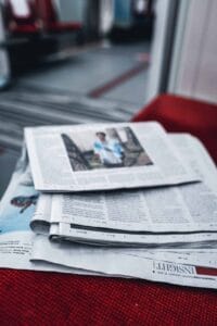 Stack of newspapers on red fabric in a public seating area, top one showing a blurred image.
