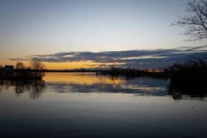 Calm water at sunset; orange and grey sky, distant city skyline, tree silhouettes framing scene.