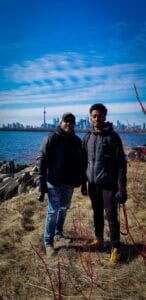 Two people in jackets stand by water with CN Tower and skyline in background, red branches nearby.