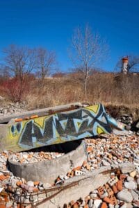 Concrete with graffiti amidst bricks, stones; dried grass, trees, lighthouse in background, blue sky.