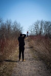 Person on dirt path photographing lighthouse amid dry grass and trees under clear sky.