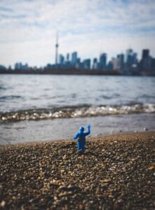Small blue toy on pebbly beach, city skyline with tower under cloudy sky in background.