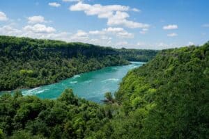 Wide turquoise river in lush forest, bordered by steep cliffs under a blue sky with clouds.