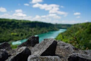 Sharp rocks in focus, river and green hills blurred in background under blue sky with clouds.