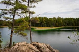 Tranquil lake with rocky shore, pine trees, green water, and distant forest under cloudy sky.