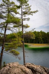Rocky outcrop with trees overlooks a lake and forest under a partly cloudy sky.